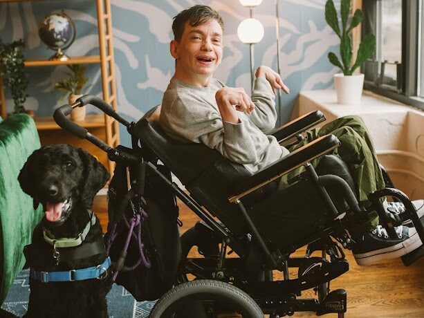 Photo of Evan and his dog, a black poodle/labrador mix named Winston. Evan is turned to the side so that you can see his wheelchair, and Winston is sitting to the left.